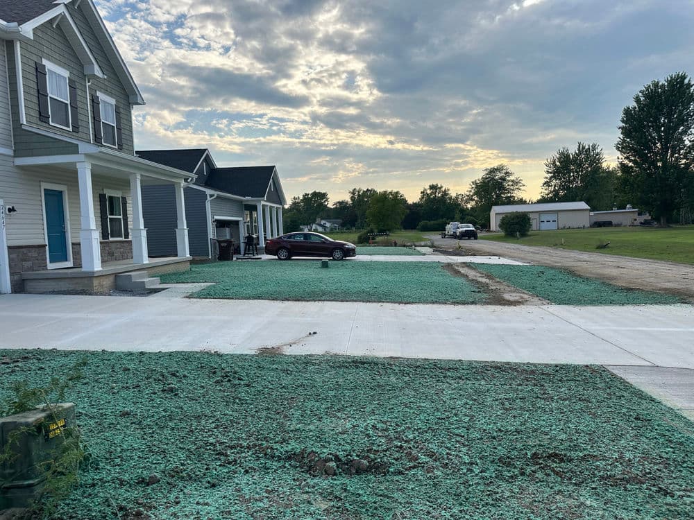 Newly paved driveways with green grass seed on a residential street under a cloudy sky.