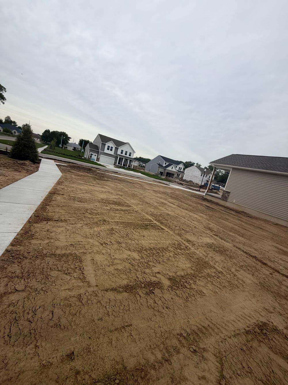 Newly graded residential lot with cleared ground, sidewalks, and nearby homes in a suburban area.