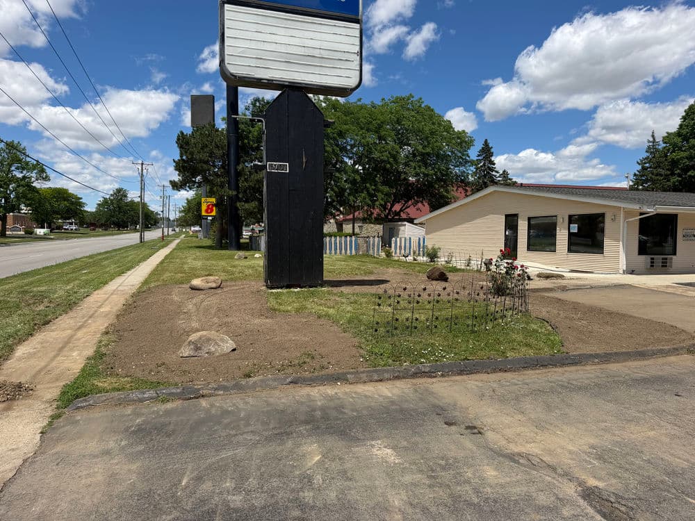 Empty lot with rocks and plants near a highway, featuring a large blank sign and a building.