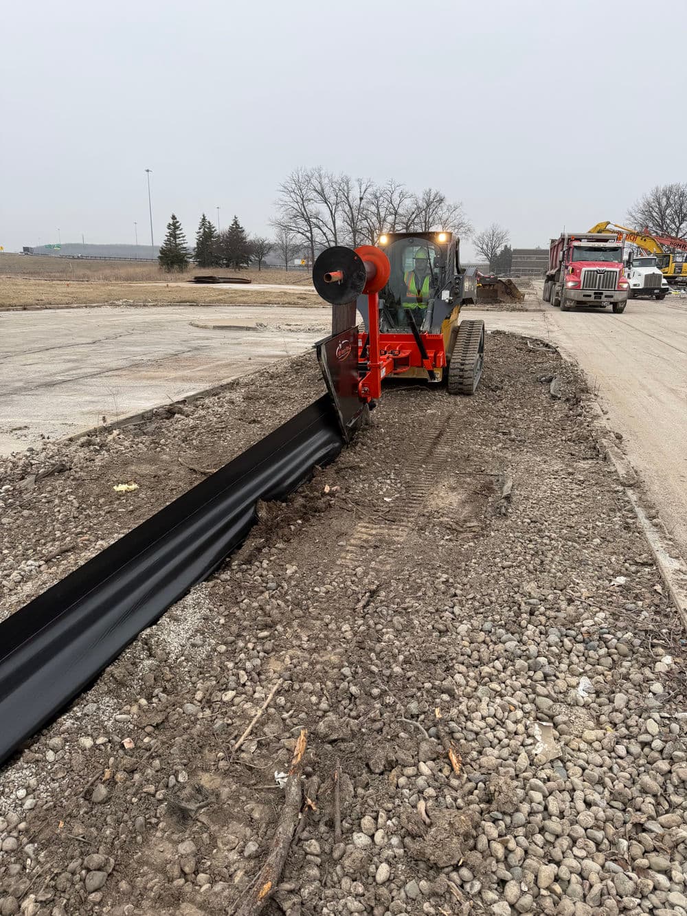 Construction site with a red machine installing black metal edging along a gravel path.