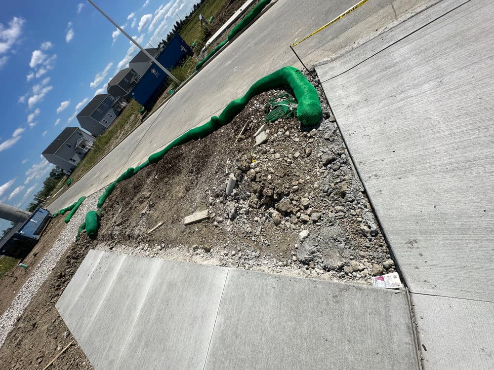 Construction site with gravel and green erosion control barriers along a concrete sidewalk.