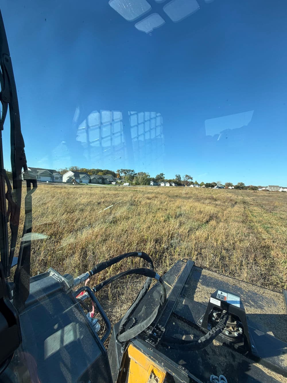 View from a tractor cab showing a field and residential homes under a clear blue sky.